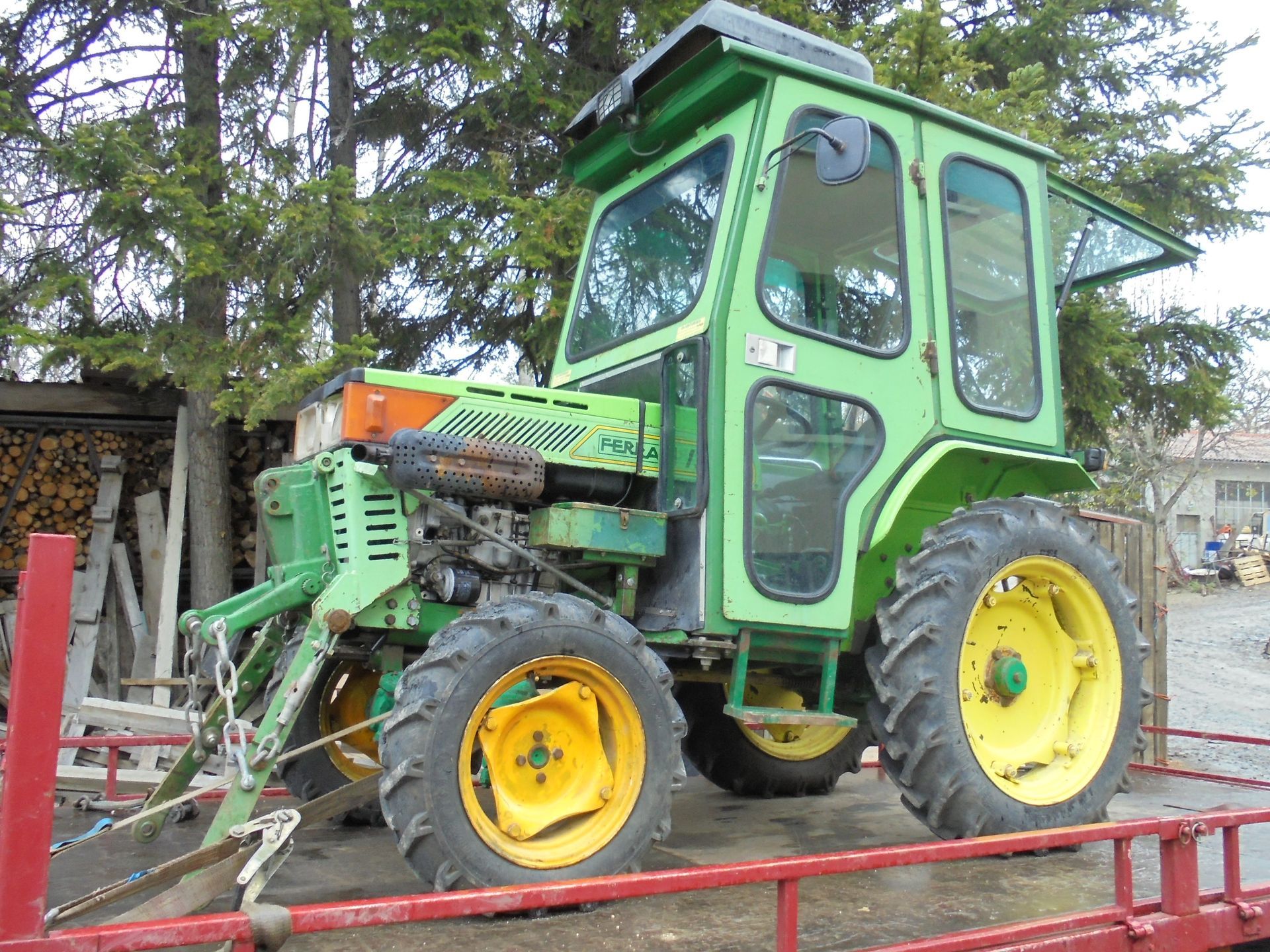 Tracteur vert à roues jaunes garé sur une remorque rouge dans une cour.