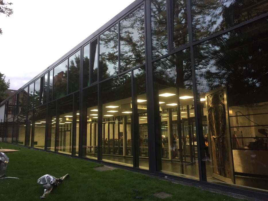 Bâtiment moderne avec de grandes baies vitrées reflétant des arbres et une pelouse verdoyante.