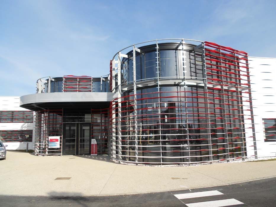 Bâtiment moderne à structure de verre et de métal rouge ; porte d'entrée visible.