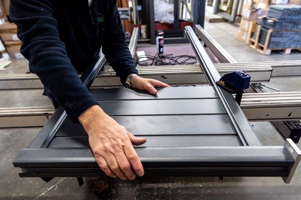 Une personne assemble un cadre métallique gris foncé, probablement pour une porte ou une fenêtre, dans un atelier.