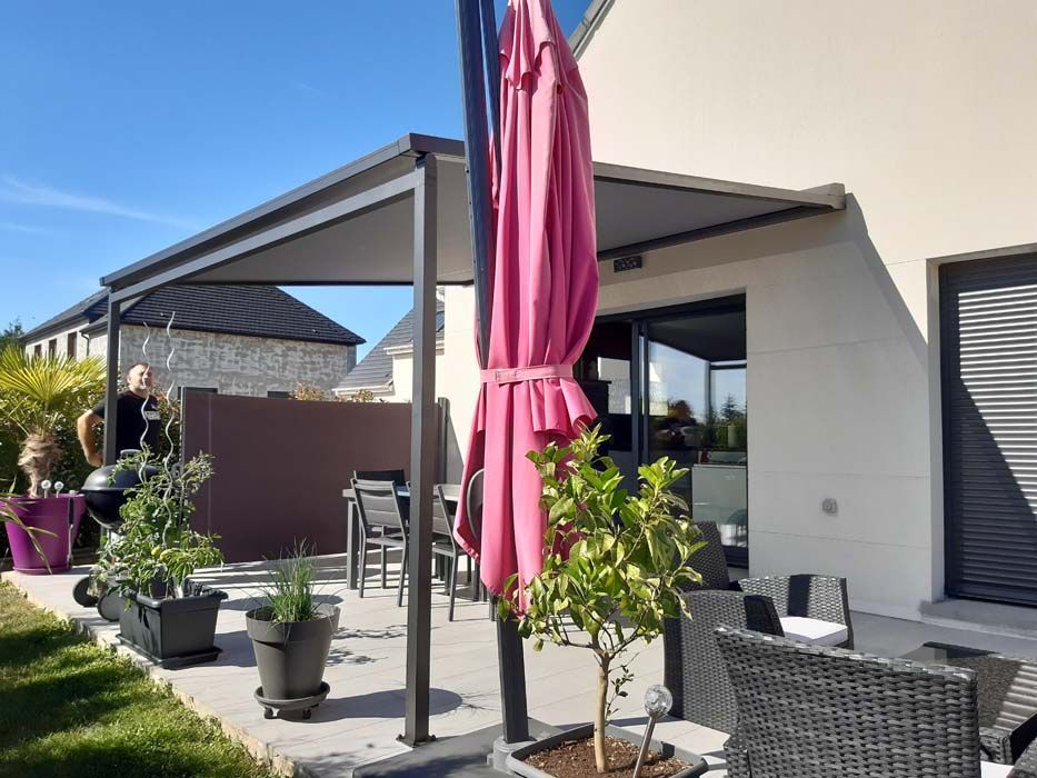 Terrasse avec pergola et parasol rose fermé, plantes en pot et mobilier de jardin sous un ciel ensoleillé.