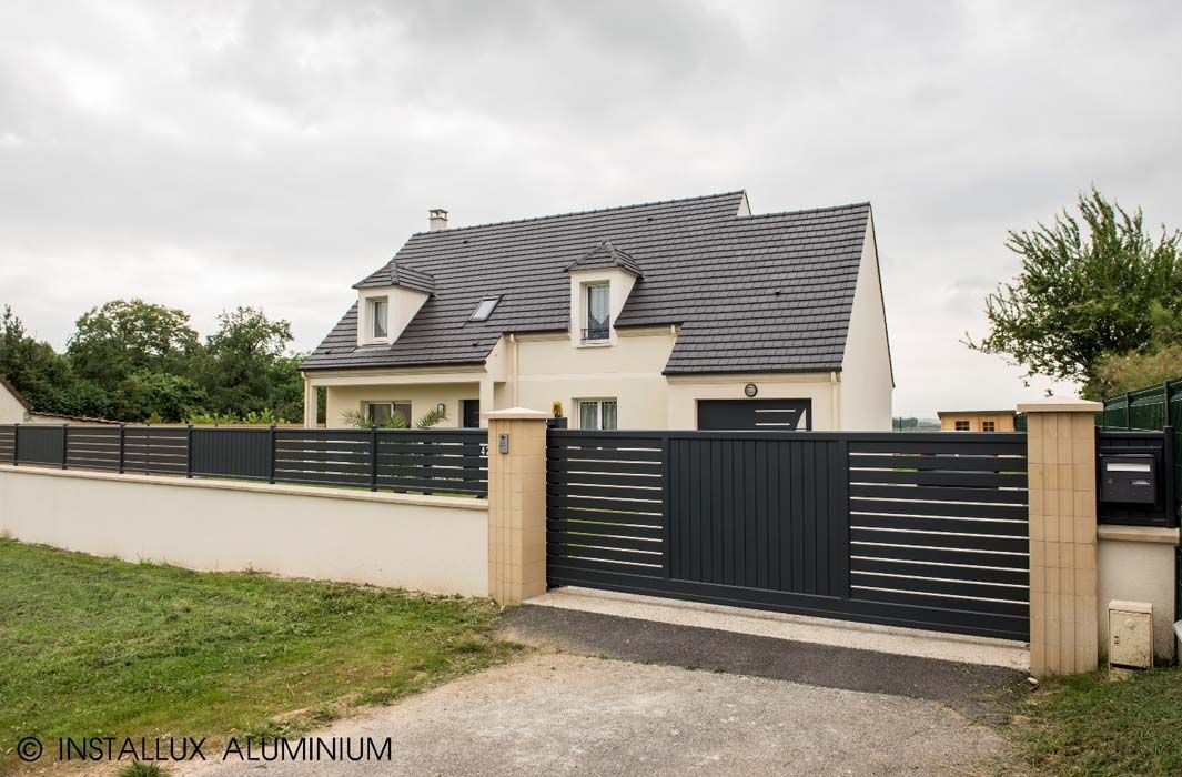 Maison grise avec un portail gris foncé, une clôture et un mur blanc, sous un ciel couvert.