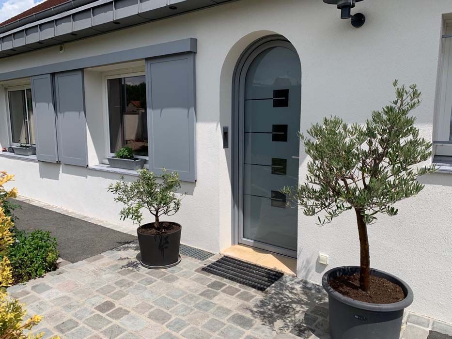 Entrée d'une maison blanche avec porte grise, volets et oliviers en pot sur une terrasse pavée.