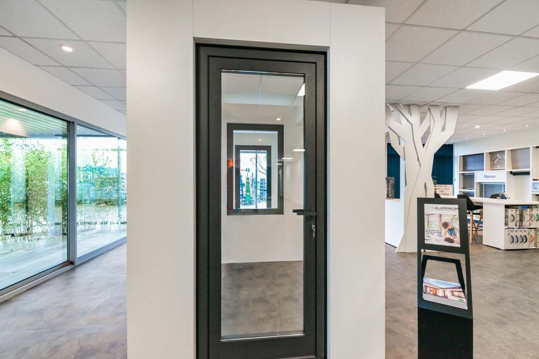 Porte vitrée à cadre sombre encastrée dans un mur blanc, donnant sur une autre porte et une fenêtre. Intérieur.