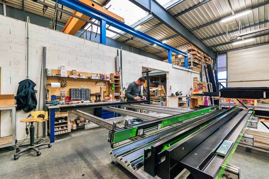Un homme travaille dans un atelier, entouré de machines et d'outils. On aperçoit des murs blancs, des poutres métalliques et des étagères.