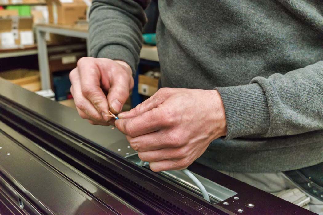 Une personne, vêtue d'une chemise grise à manches longues, relie des fils électriques à un rail métallique. Elle se trouve à l'intérieur.