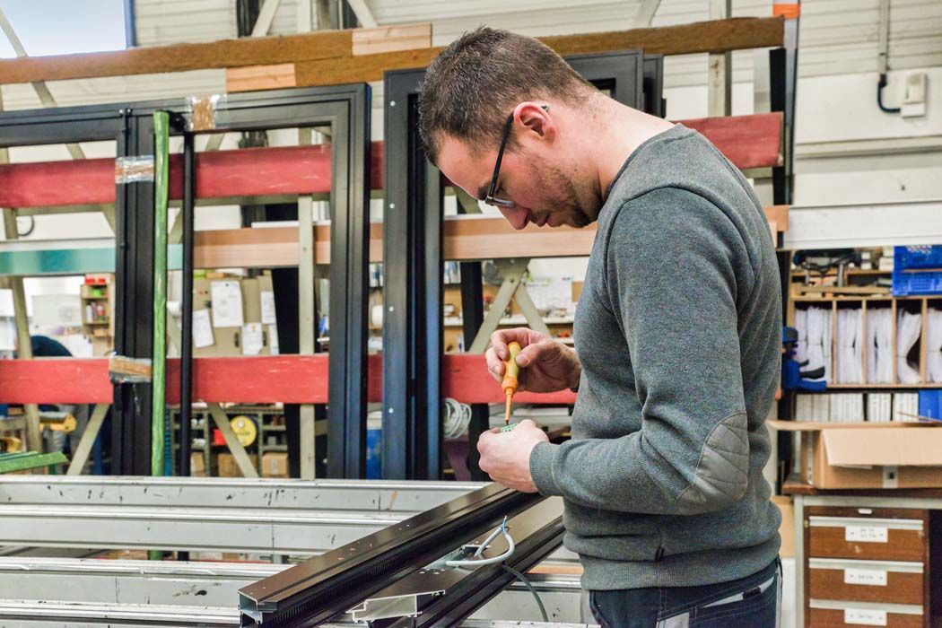 Un homme portant des lunettes utilise un tournevis pour assembler un cadre de fenêtre dans une usine.