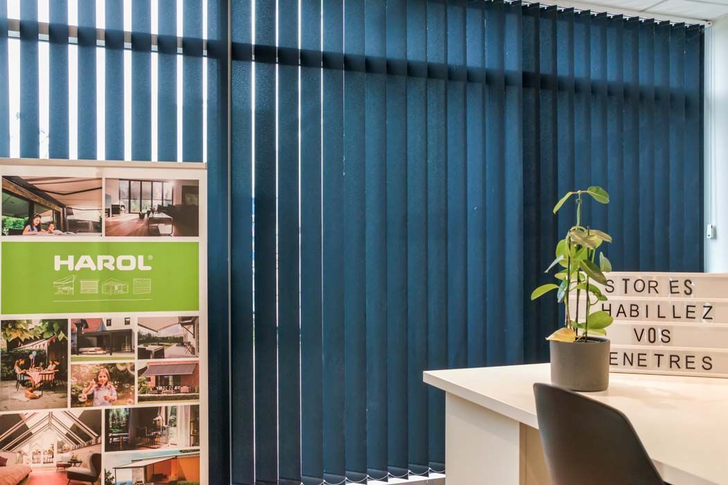 Stores verticaux bleus dans une pièce lumineuse avec un bureau, une pancarte et une plante.