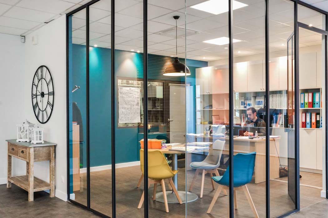 Bureau aux parois de verre avec un mur d'accent turquoise, table ronde, chaises colorées et une personne travaillant à un bureau.