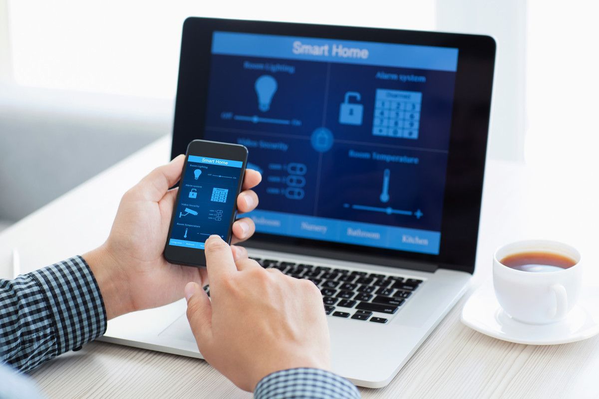 Personne utilisant un smartphone et un ordinateur portable pour contrôler des appareils domotiques, notamment l'éclairage, la sécurité et la température.