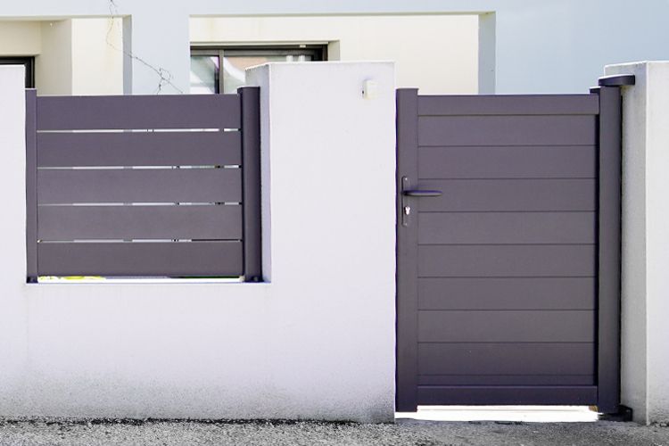 Portail et section de clôture à lattes horizontales grises encastrés dans des murs en béton blanc.
