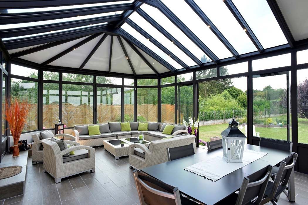 Véranda avec toit et murs en verre, meublée d'un coin salon et d'une table à manger donnant sur un jardin.