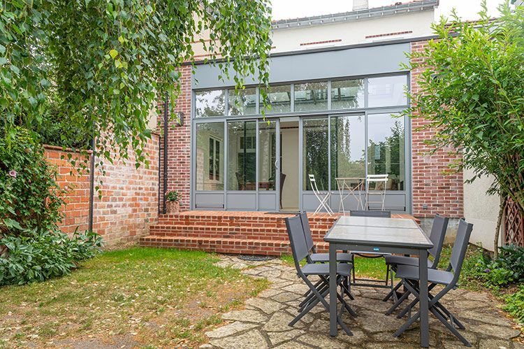 Jardin avec terrasse en briques et dépendance vitrée. Table et chaises grises sur la pelouse. Arbres et feuillage verts.