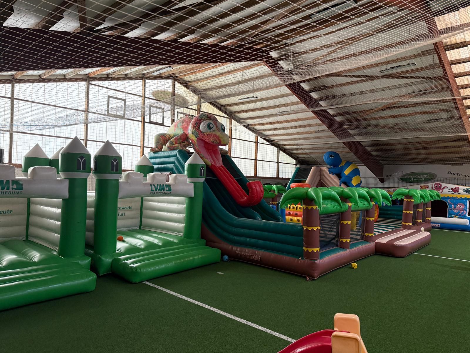 Indoor-Spielplatz mit aufblasbaren Burgen, Rutschen und Palmen auf grünem Boden.