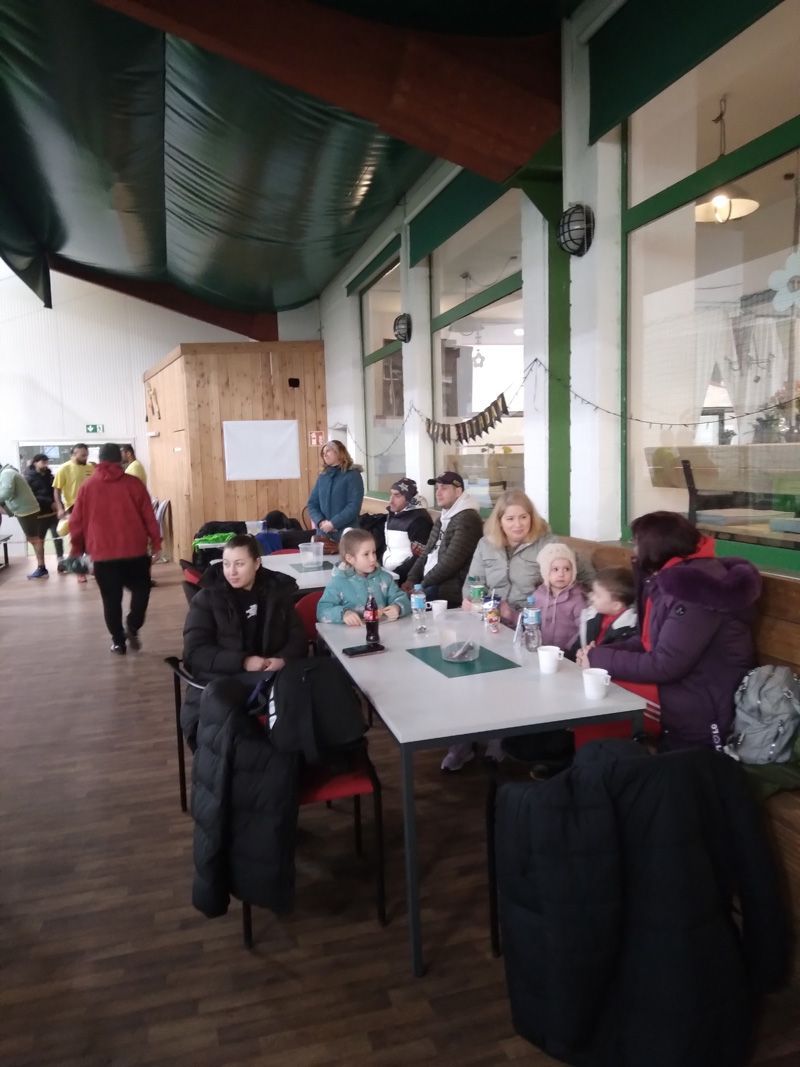Menschen an Tischen in einem Gebäude, möglicherweise einem Café. Eine Person in einer roten Jacke geht vorbei.