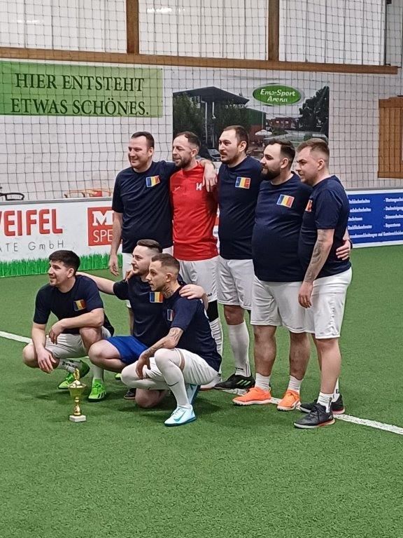 Fußballmannschaft posiert mit Trophäe in der Halle; Männer in blau-weißen Uniformen, einer in roten, auf grünem Rasen.