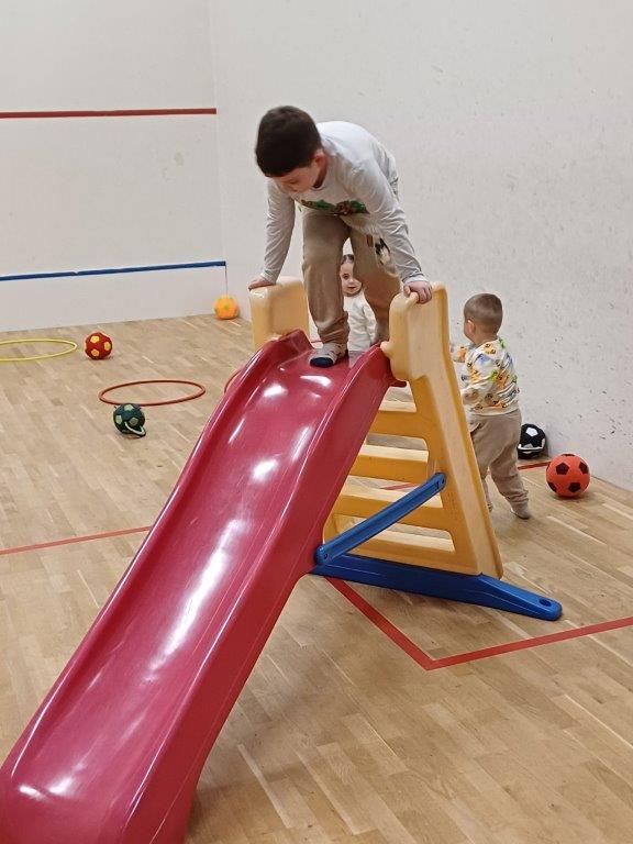 Junge klettert auf Rutsche, zwei Kleinkinder schauen zu. Rote Rutsche im Raum mit Bällen.