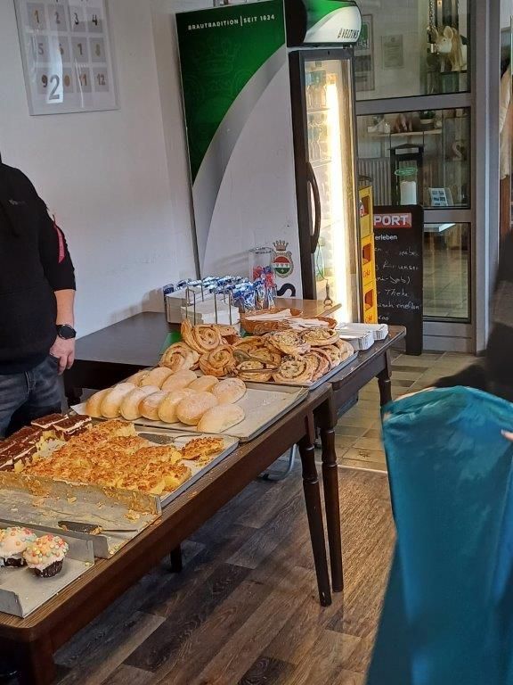 Ein mit Backwaren, Getränken und einer Kühlbox beladener Tisch in einem Geschäft. Eine Person steht in der Nähe.