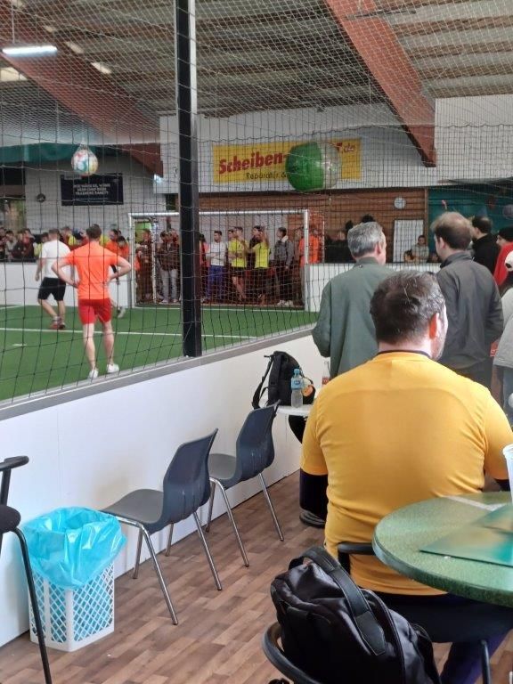 Hallenfußballspiel: Spieler in Orange und Schwarz, Zuschauer, Ball in der Luft, Tor im Hintergrund.
