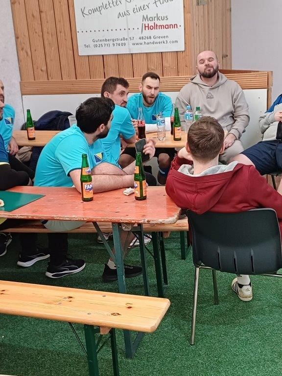 Eine Gruppe von Männern sitzt in einem Raum um einen Tisch mit Bierflaschen. Einige tragen blaugrüne Hemden und unterhalten sich.
