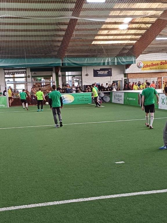 Hallenfußballspiel: Spieler in grün-schwarzen Trikots auf grünem Rasen. Zuschauer beobachten das Spiel von der Seitenlinie.