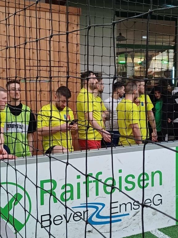 Männer in gelben Trikots schauen hinter einem Tor ein Fußballspiel an. Raiffeisen-Schild sichtbar.
