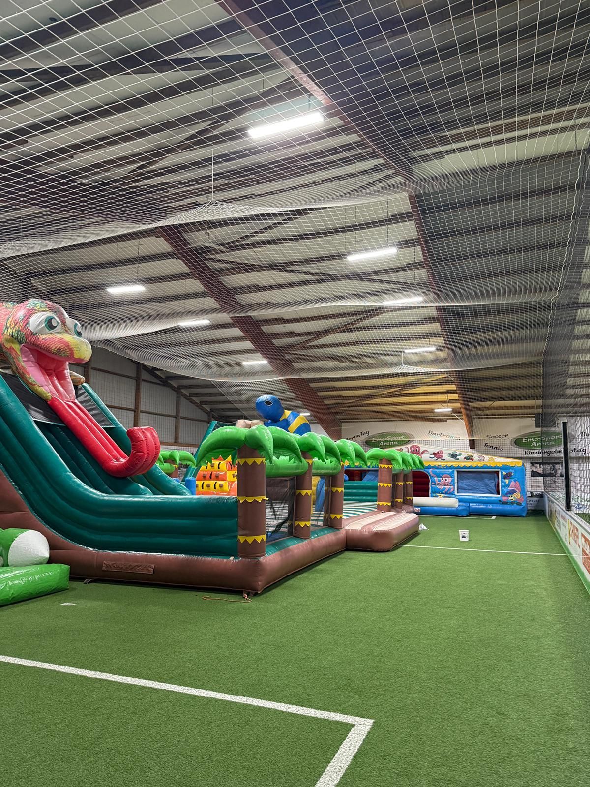 Indoor-Spielplatz mit aufblasbaren Spielgeräten auf grünem Rasen unter einem Netz.