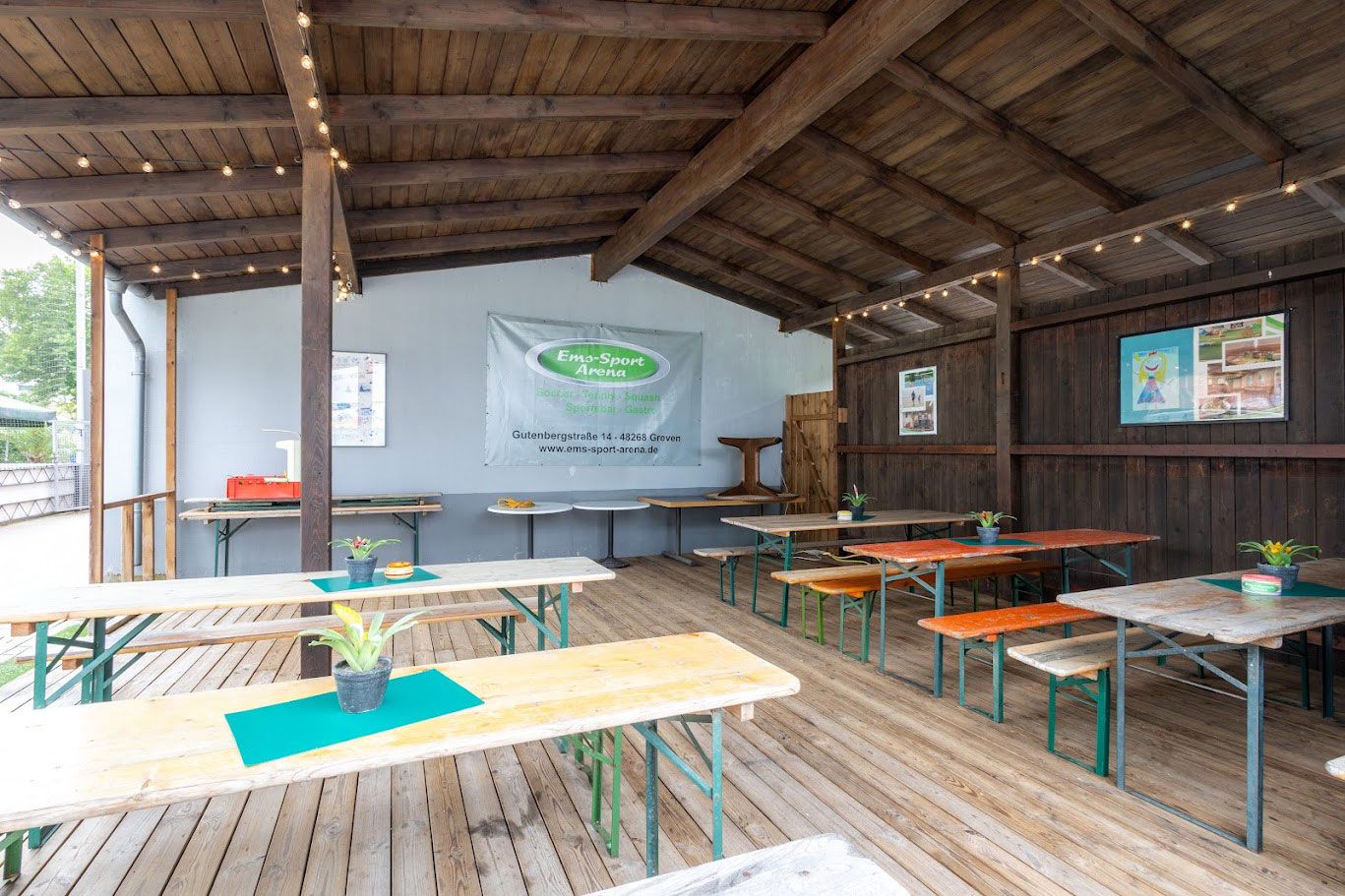 Überdachter Sitzbereich im Freien mit Picknicktischen, einem Banner und Lichterketten.