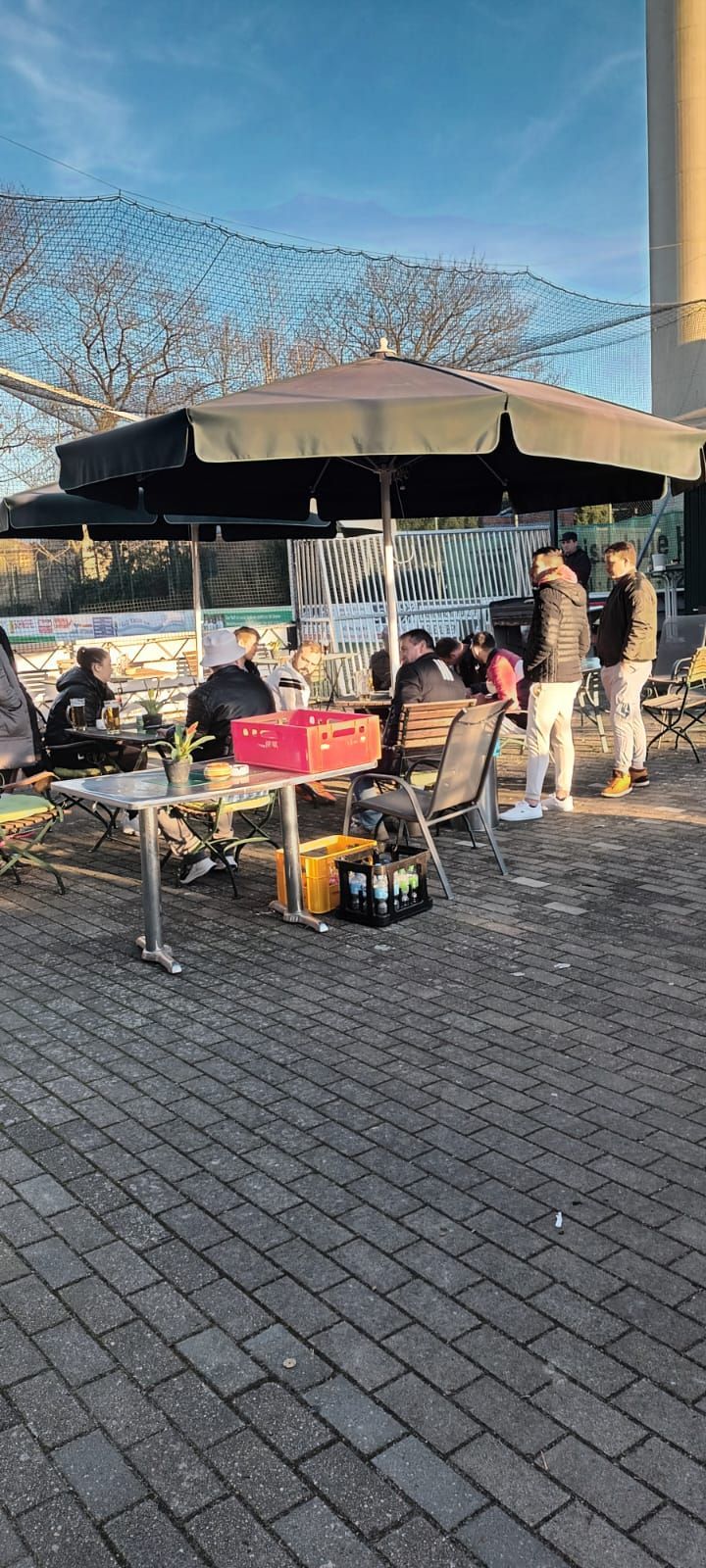 Eine Gruppe von Menschen sitzt unter einem Sonnenschirm an Tischen auf einem gepflasterten Gehweg.