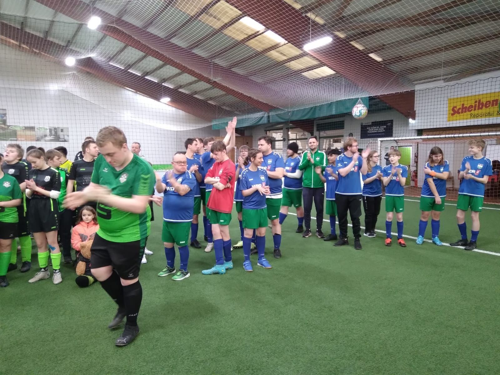 Fußballmannschaften in grünen und blauen Uniformen feiern in einem Hallenstadion, klatschen und lächeln.