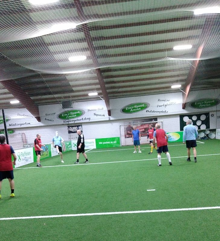Hallenfußballspiel; Spieler in verschiedenfarbigen Trikots auf grünem Rasen, unter Netzen und Deckenbeleuchtung.