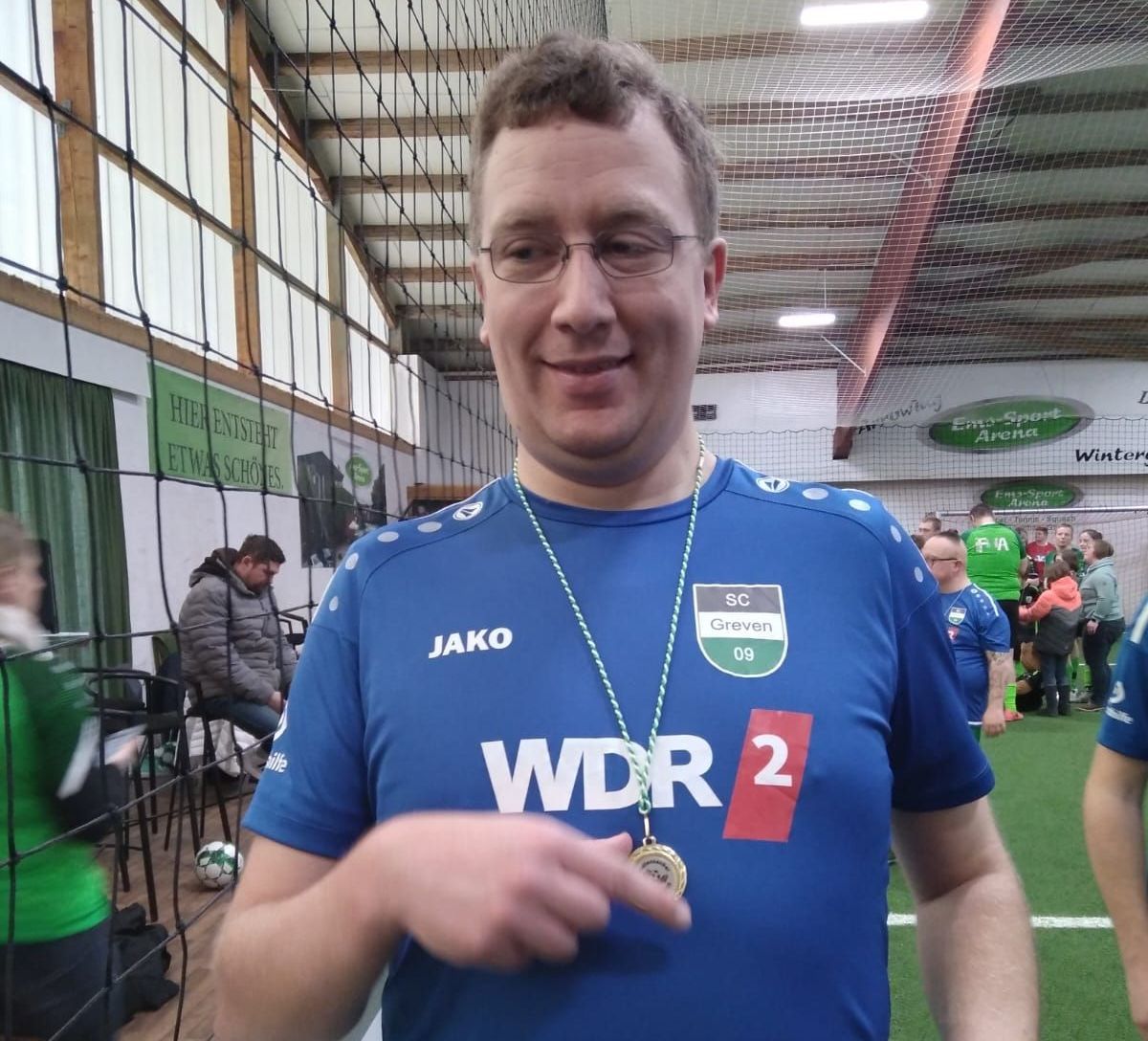 Mann im blauen Fußballtrikot mit Medaille lächelt bei einer Veranstaltung im Innenbereich.