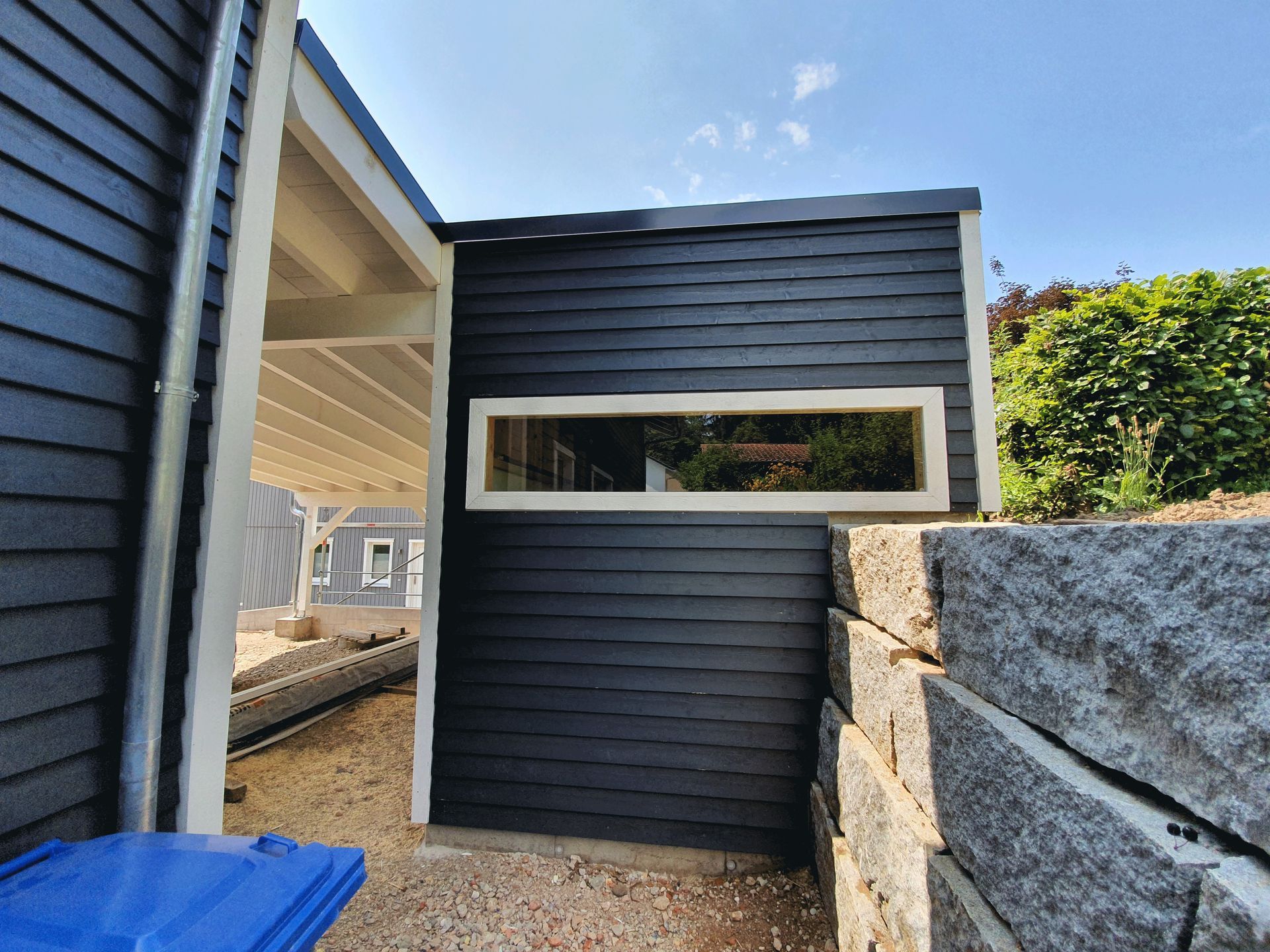 Ein schwarzes Gebäude mit einem Fenster und einer blauen Mülltonne davor.