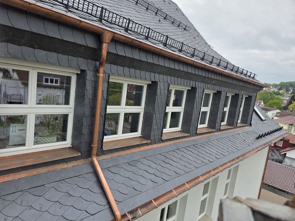Dunkles Schieferdach mit Kupferdachrinnen und mehreren weiß gerahmten Fenstern an der Außenseite eines Gebäudes.