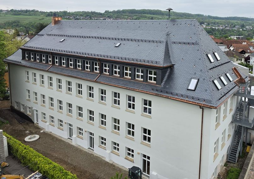Dreistöckiges weißes Gebäude mit dunkelgrauem Schieferdach und zahlreichen Fenstern, in einer Stadt an einem bewölkten Tag.