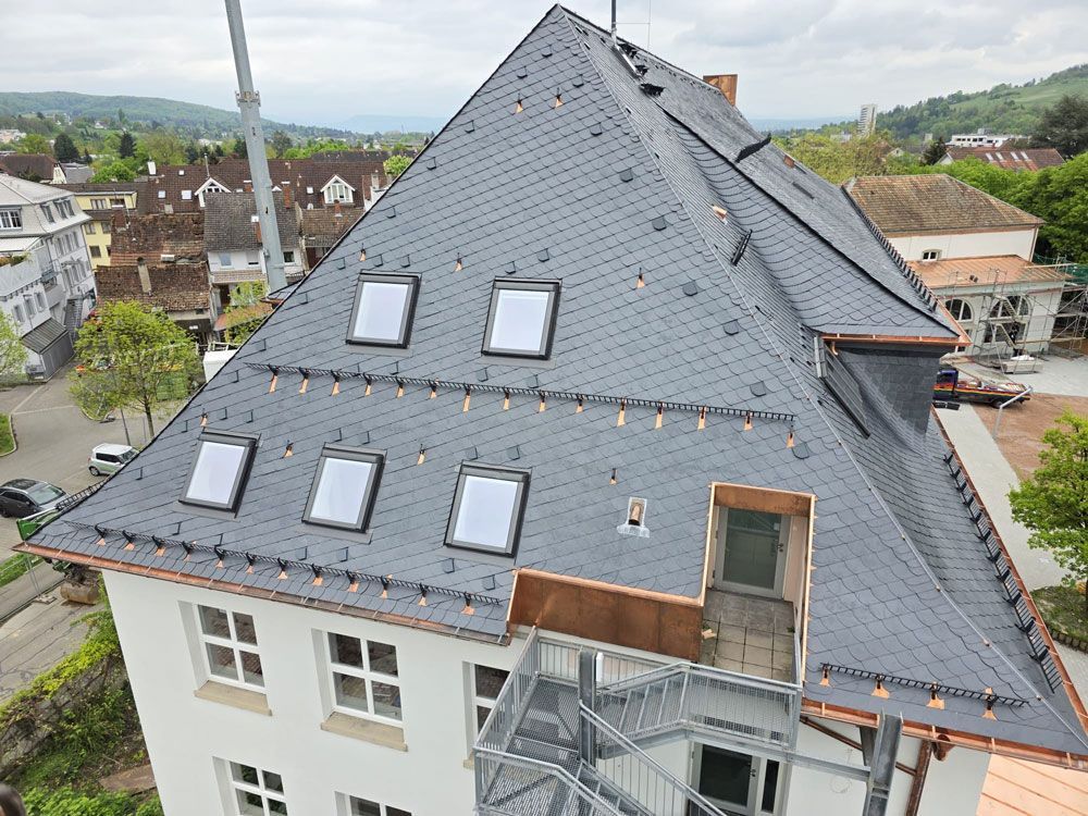 Weißes Gebäude mit dunkelgrauem Schieferdach, Kupferverkleidung, mehreren Oberlichtern und metallener Feuerleiter.