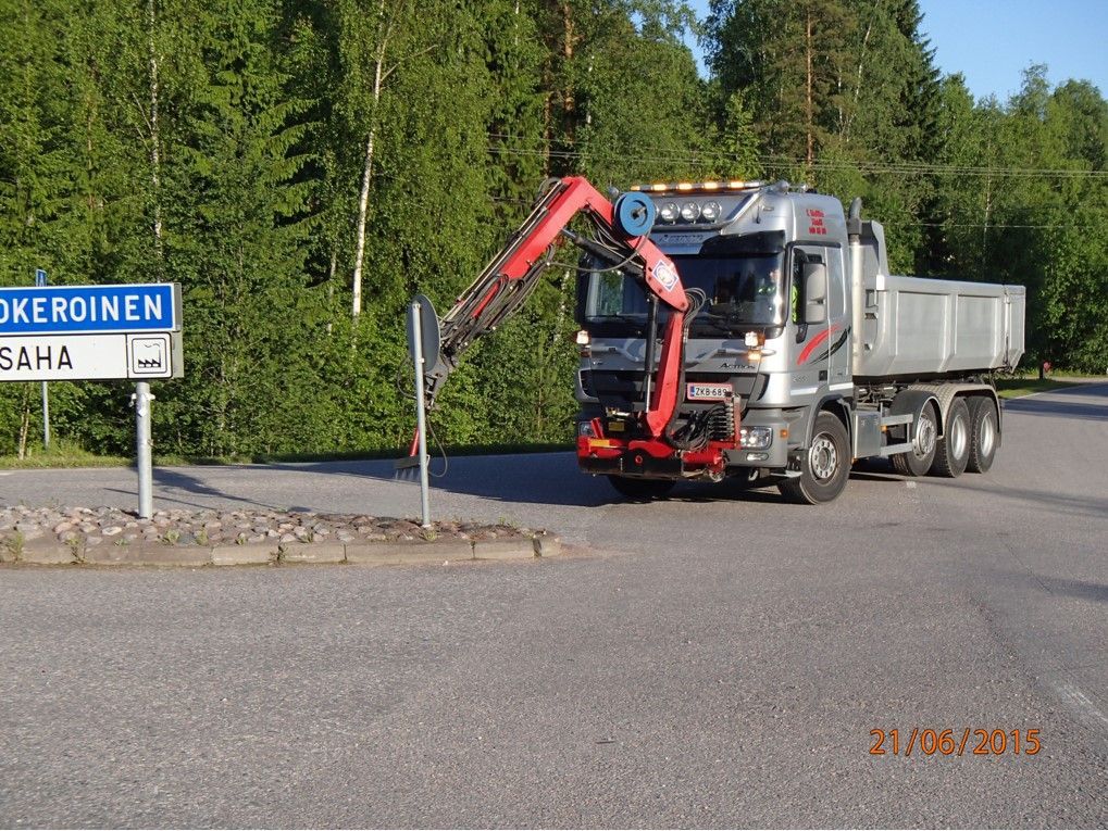 Punainen kuorma-auto nosturilla pysäköitynä tien varteen kyltin ja puiden viereen