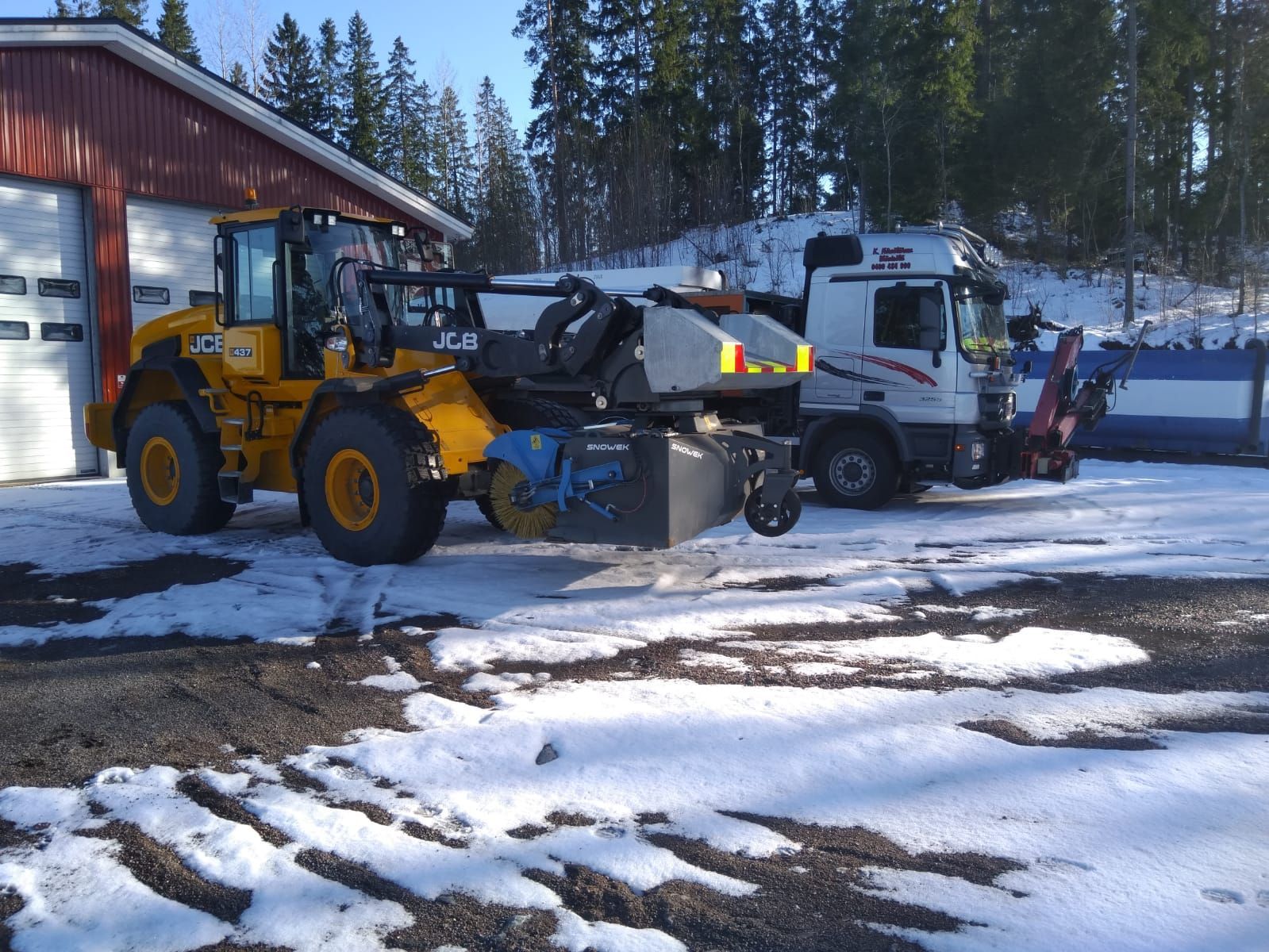 Keltainen etukuormaaja ja valkoinen kuorma-auto pysäköityinä punaisen rakennuksen ulkopuolelle lumisella pihalla.