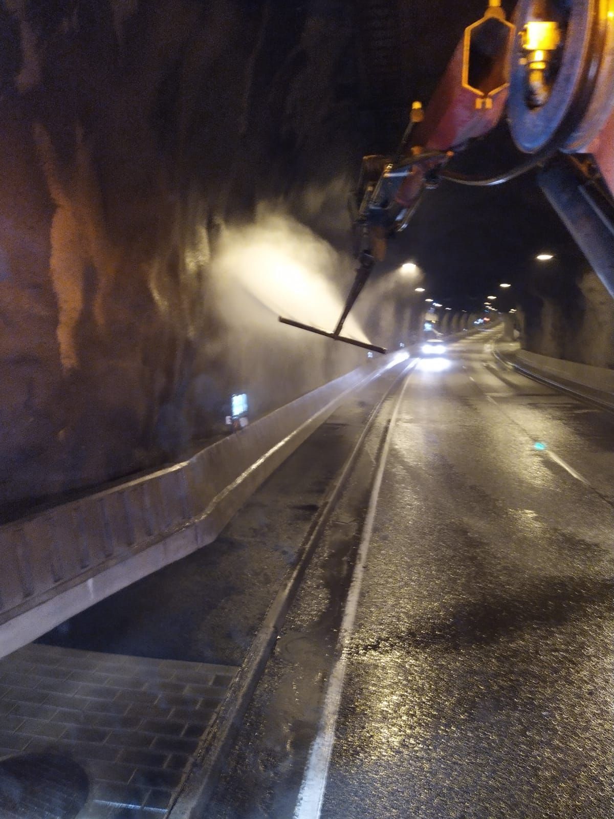 Työntekijä suihkuttaa vettä hämärässä tunnelissa märän tien ja ajovalojen ollessa edessä