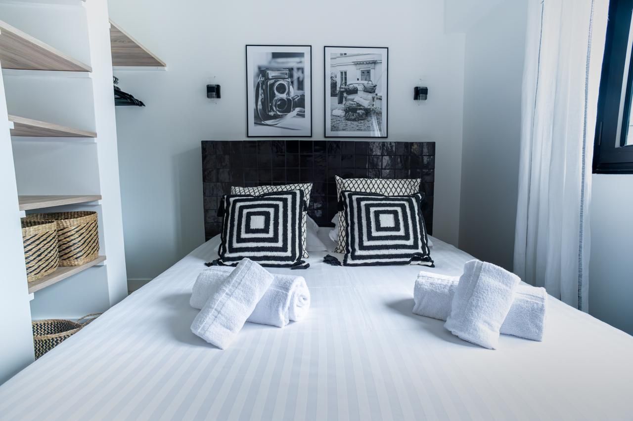 Chambre lumineuse et blanche avec un lit soigneusement fait, des coussins noirs et blancs, des œuvres murales et des étagères près de la fenêtre