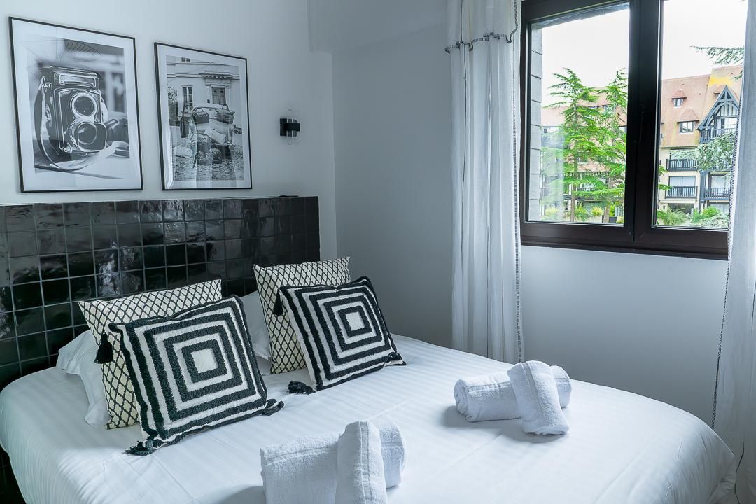 Chambre lumineuse avec un lit blanc, des coussins noirs et blancs, des œuvres encadrées et une fenêtre avec des rideaux