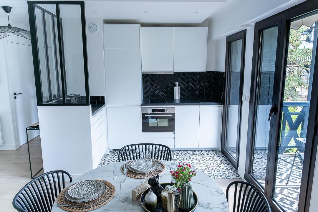 Cuisine lumineuse et moderne avec des placards blancs, des touches de noir, une table ronde et des fenêtres ouvertes sur un balcon