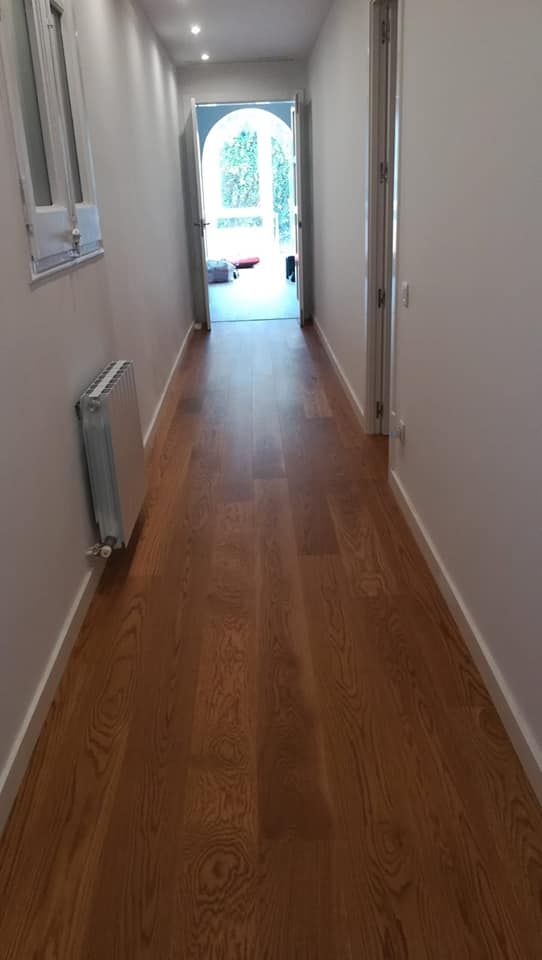 Pasillo largo con suelo de madera, paredes blancas y puerta al final con luz exterior.