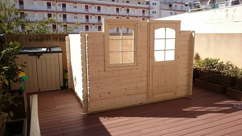 Casa de juegos de madera sin terminar con ventana y puerta, instalada en una terraza con otras estructuras de madera.