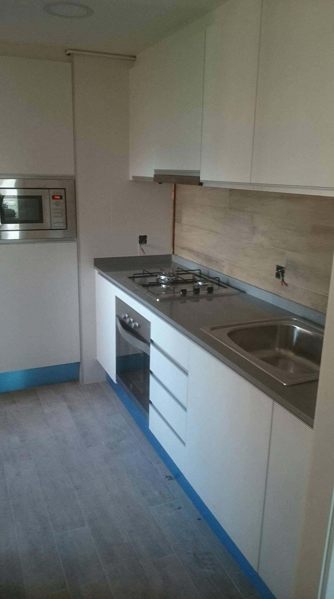 Gabinetes y encimeras de cocina blancos, electrodomésticos grises y protector contra salpicaduras con apariencia de madera en una habitación luminosa.