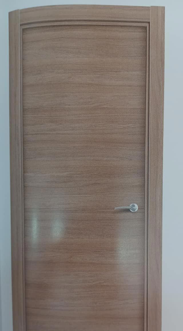 Puerta de madera clara con tirador plateado y marco sencillo sobre una pared blanca.