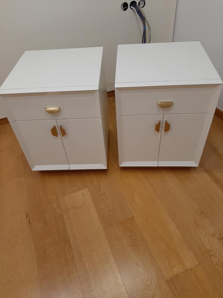 Dos mesitas de noche blancas con tiradores dorados sobre un suelo de madera.