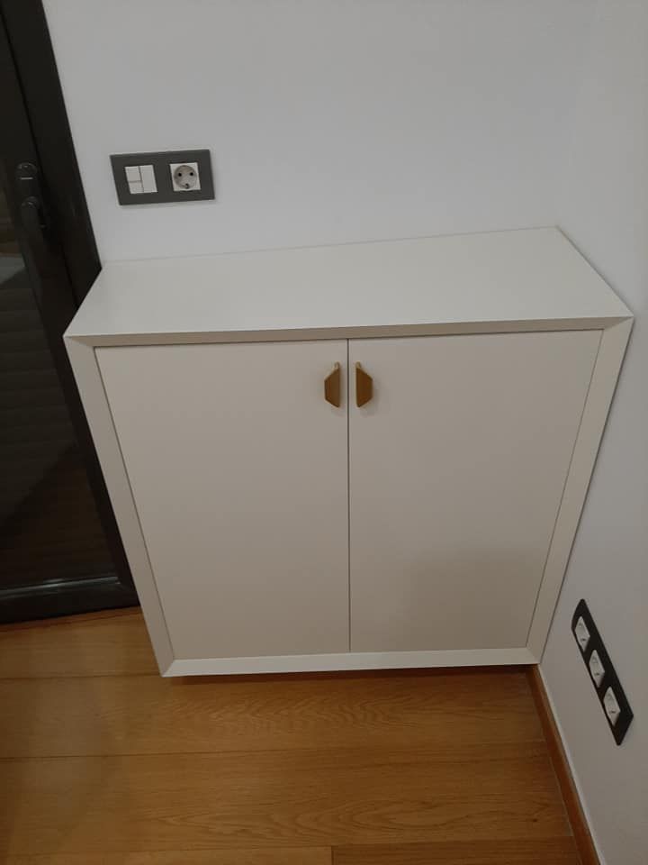 Armario blanco con tiradores dorados, contra una pared blanca con enchufes. Suelo de madera.