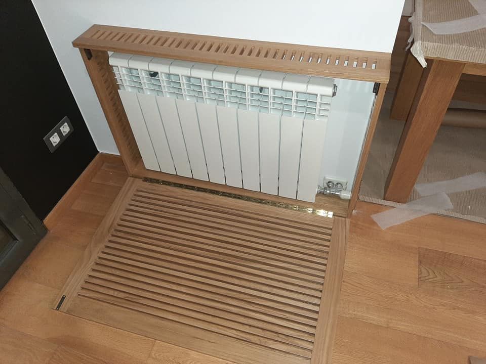 Cubierta de radiador de madera, abierta, revelando un radiador blanco contra una pared blanca y un piso de madera.