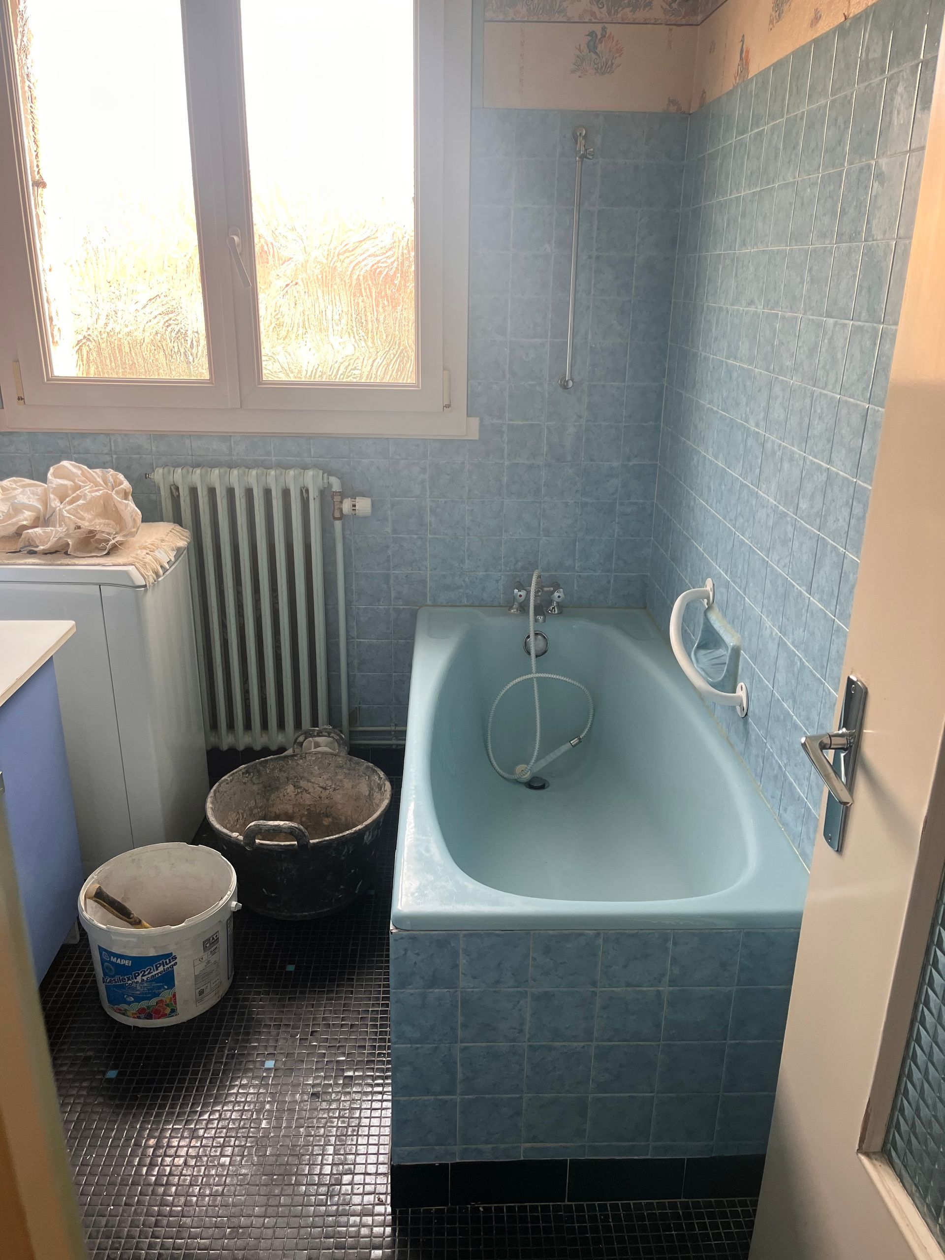 Salle de bains carrelée de bleu avec baignoire et fenêtre.